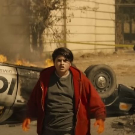 Julian Dennison has fire hands as there is a police car upside down behind him.